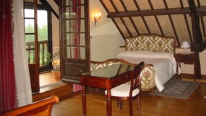 Une chambre avec un plafond en bois, un lit recouvert d’une literie à motifs floraux, une chaise en bois, une table et une fenêtre avec des rideaux.