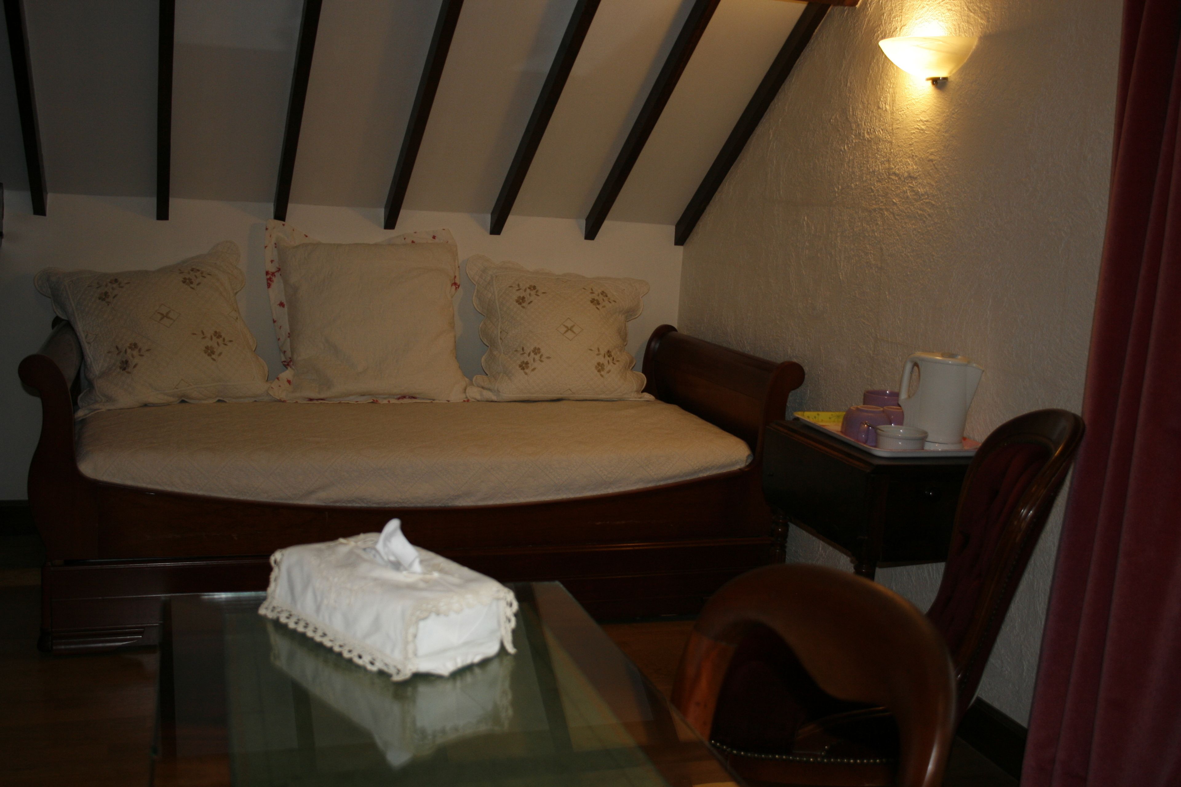 A bedroom with a bed, a bedside table with a lamp, a chair, and a glass table with a tissue box.