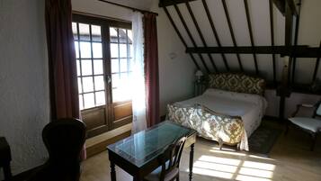 A room with a wooden table and chairs, a bed with floral bedding, and a window with curtains.