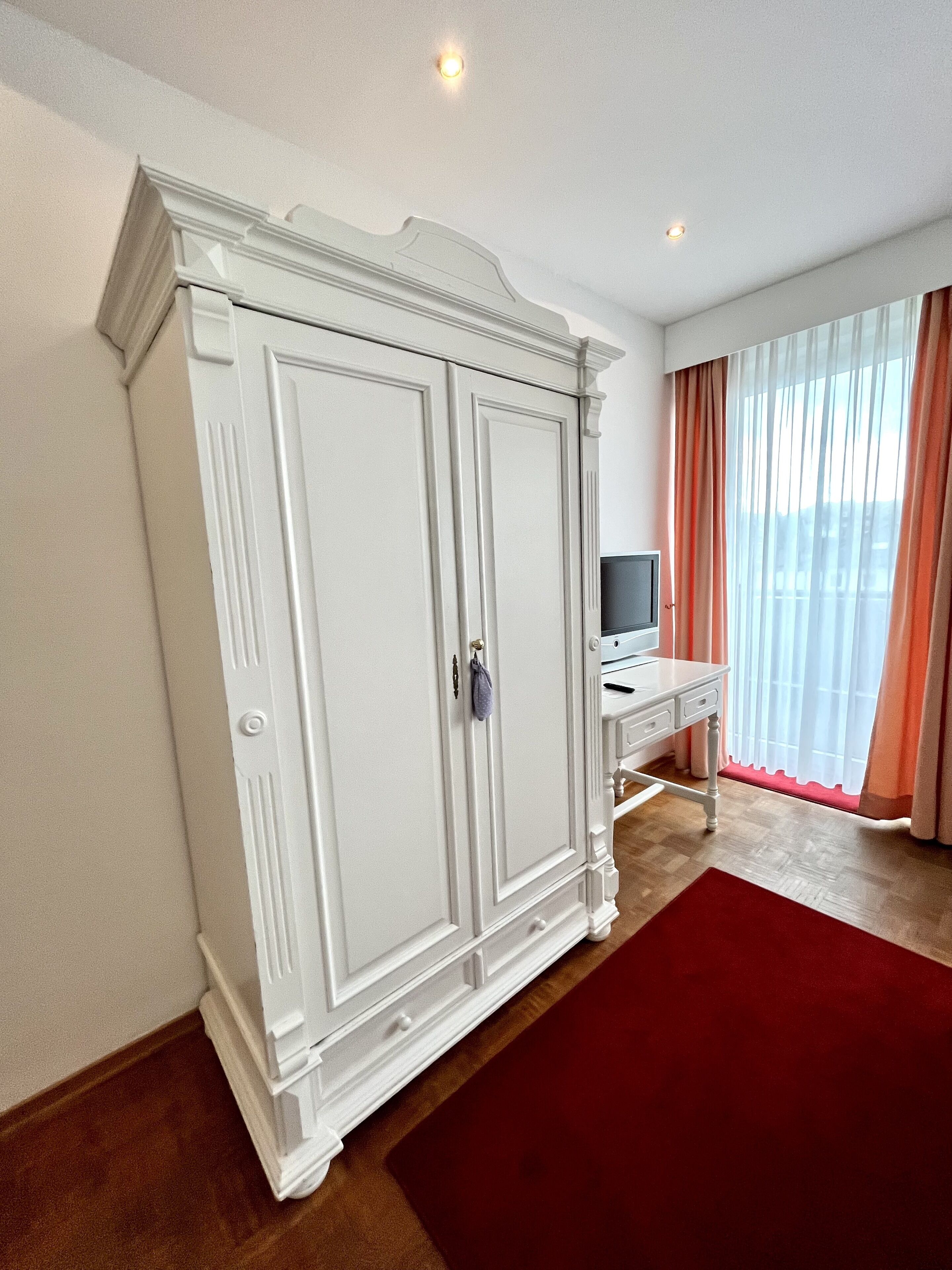 Una habitación con un armario blanco grande, un escritorio pequeño con televisor y una alfombra roja.