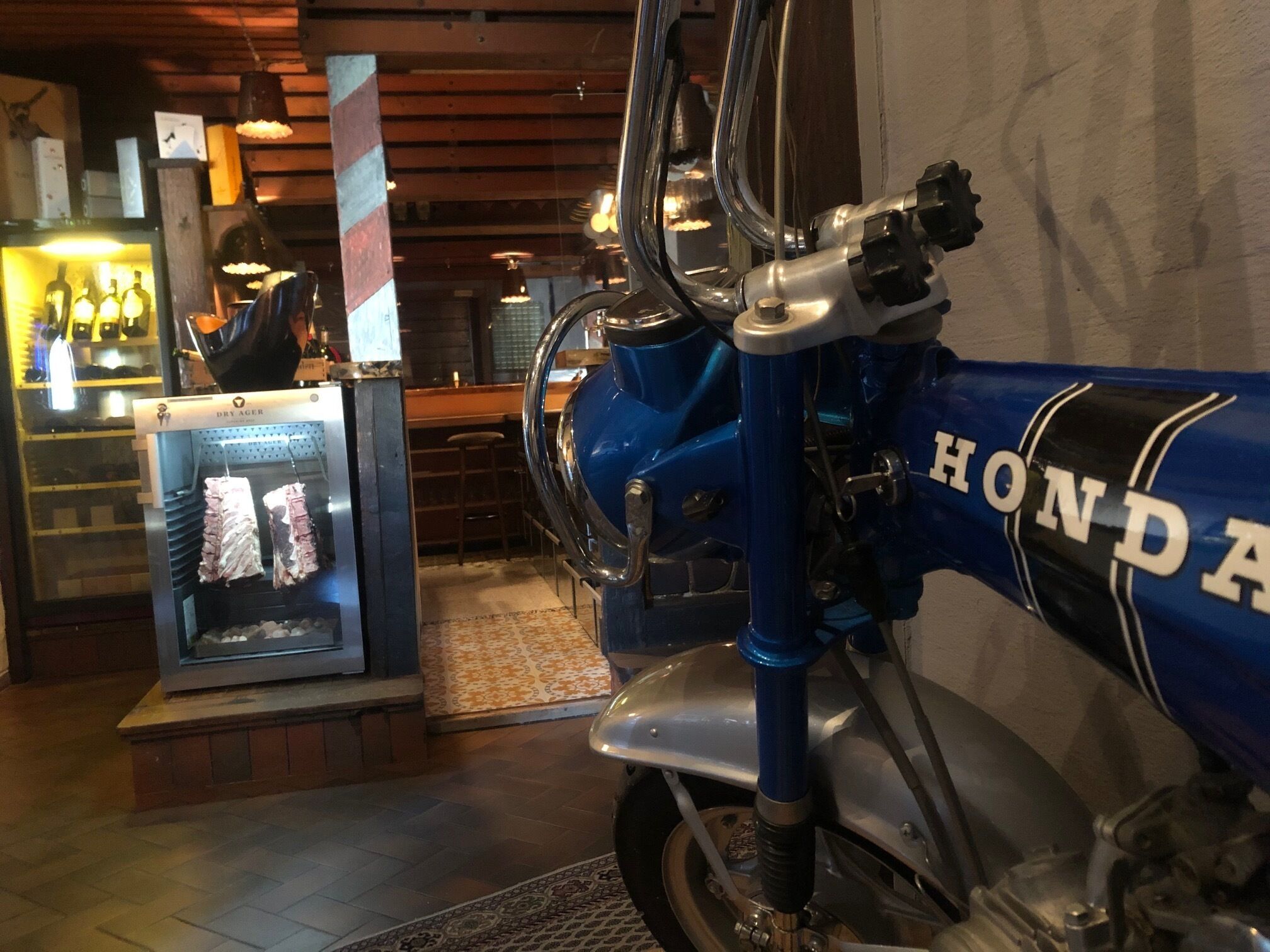 A motorcycle with a blue and silver color scheme, a visible "HONDA" logo, and a bar area with stools and a refrigerator in the background.