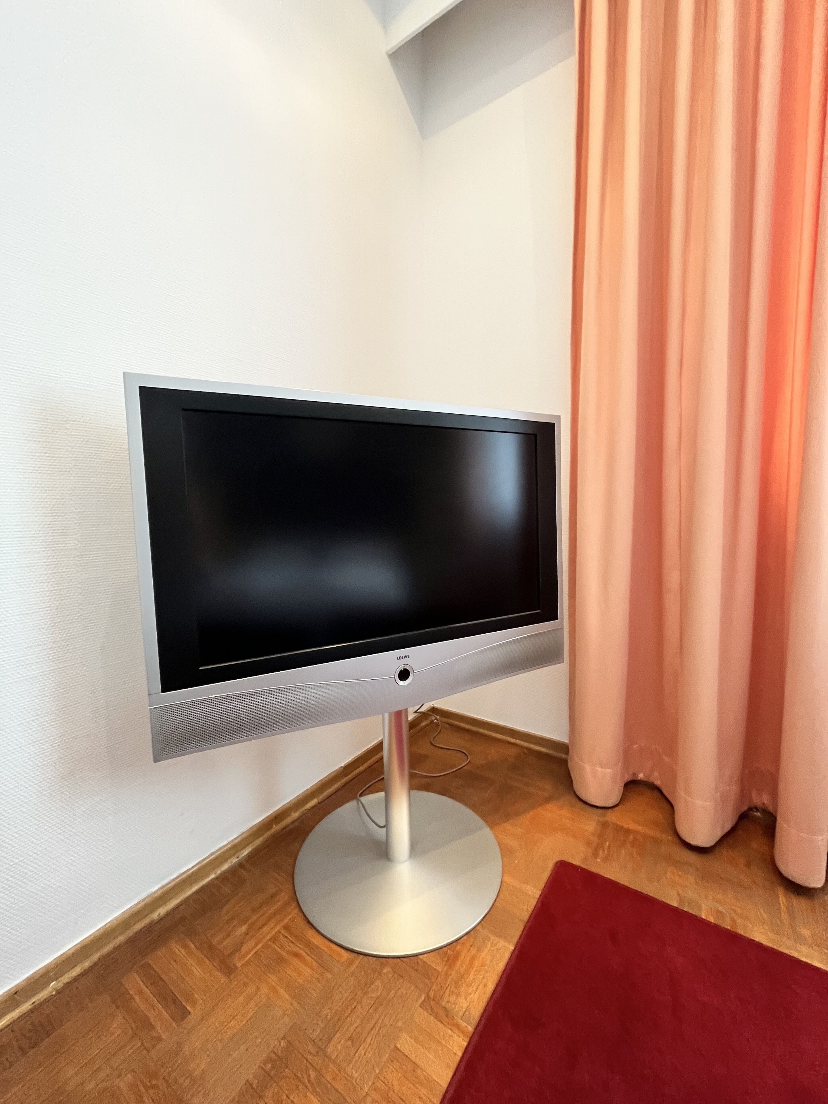 Un téléviseur à écran plat, posé sur un meuble, dans une pièce dont le sol est en bois et où se trouve un tapis rouge.