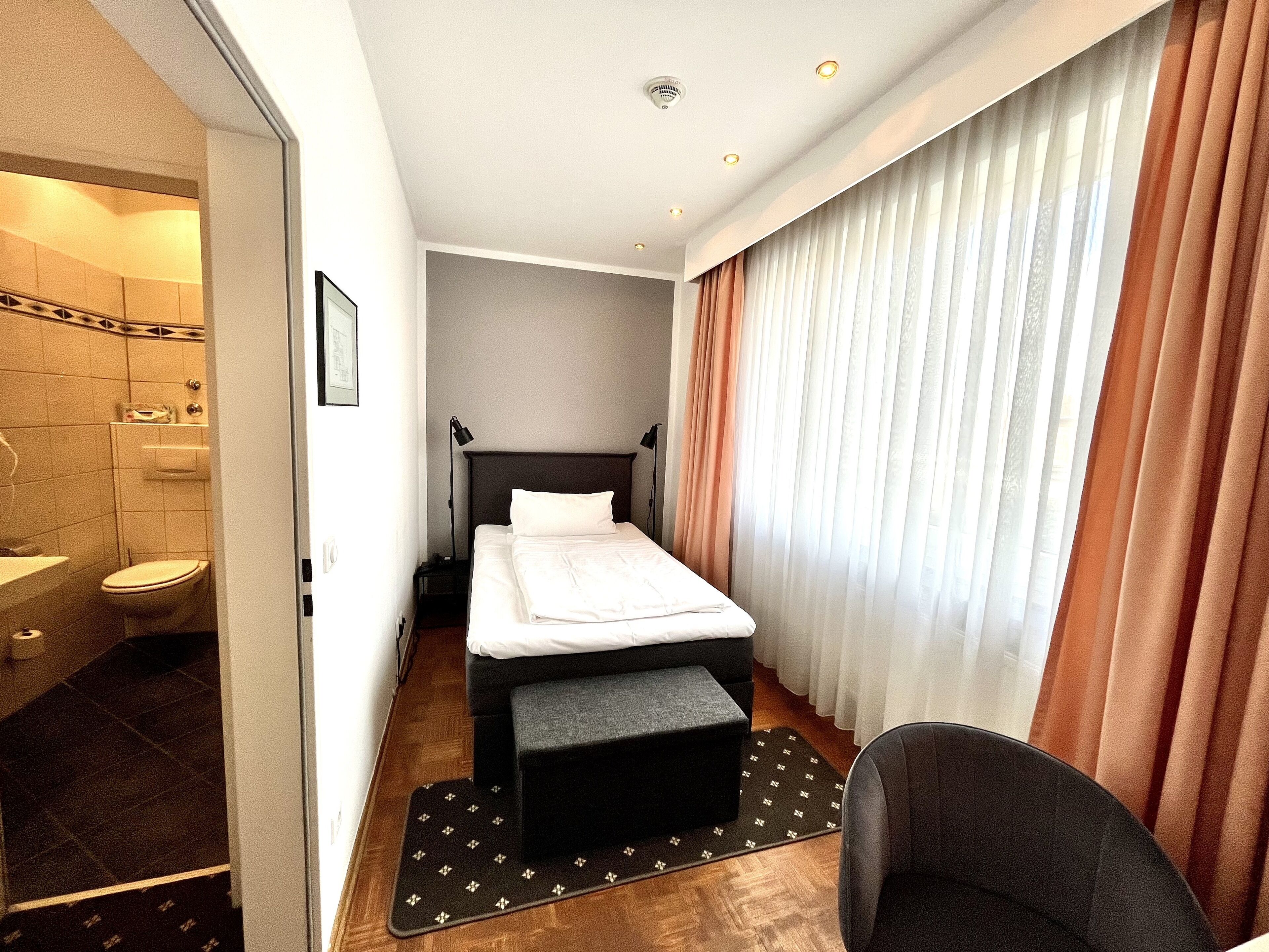 A hotel room with a bed, a chair, a small table, a bathroom with a toilet and sink, and a window with curtains.