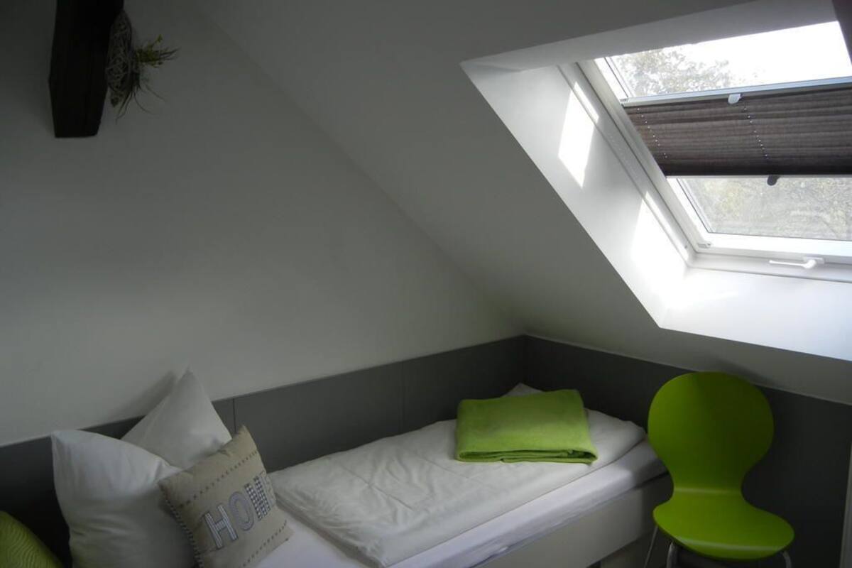 A small, single-bed room with a skylight, a green chair, and a pillow with "HO" on it.