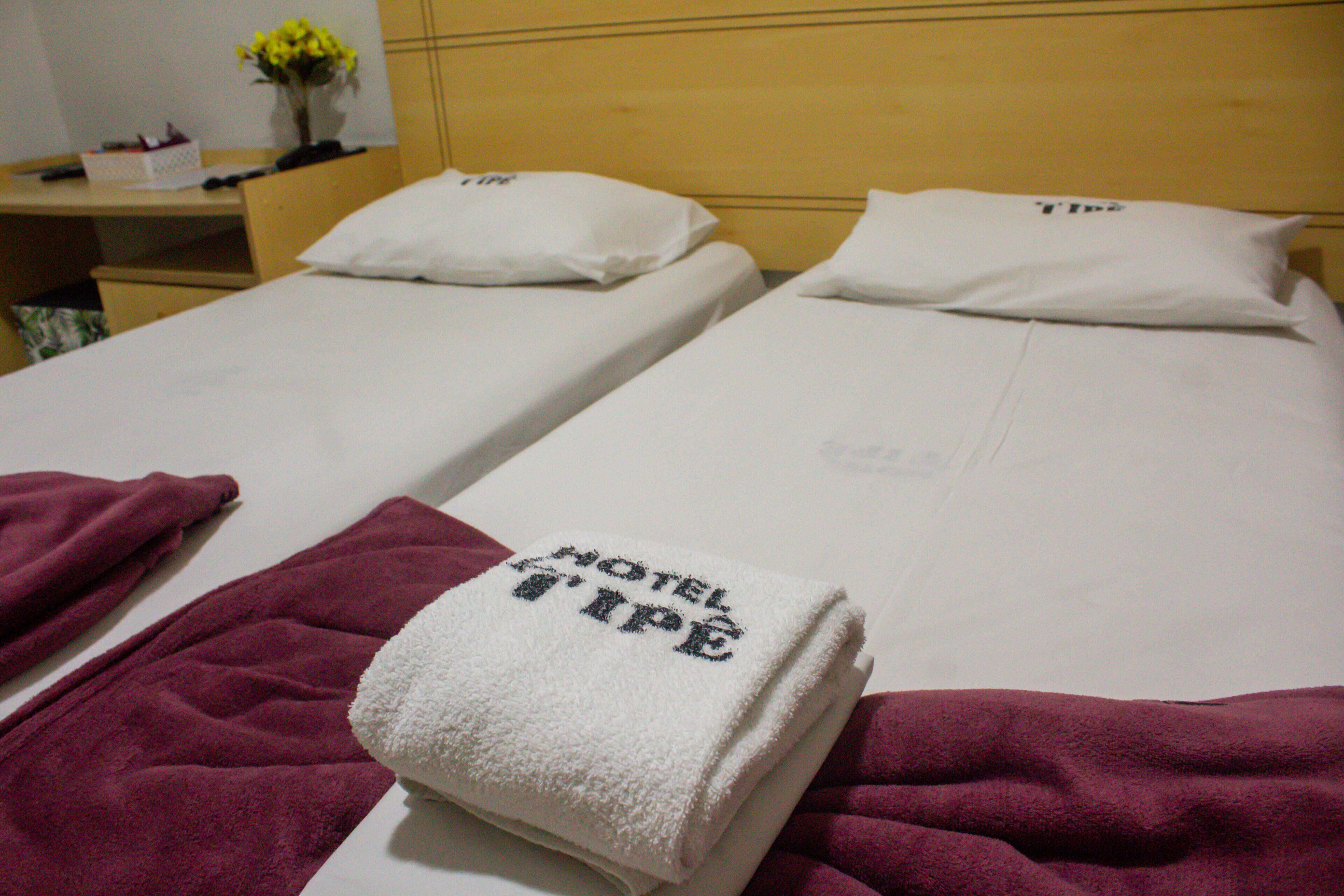 Dos camas individuales con sábanas y almohadas blancas, un cabecero de madera y una mesita de noche con un jarrón de flores amarillas.