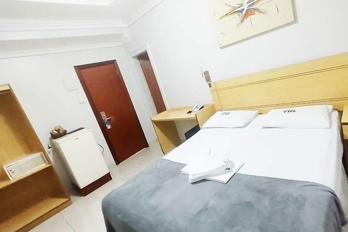 A hotel room with a bed, a desk, a chair, a small refrigerator, and a door.