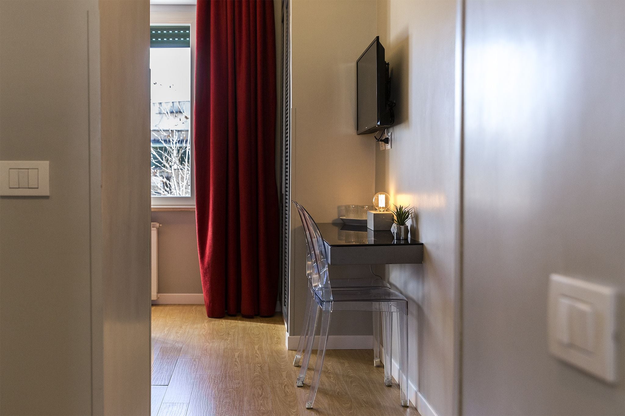 A room with a desk, a chair, a television mounted on the wall, and a window with red curtains.