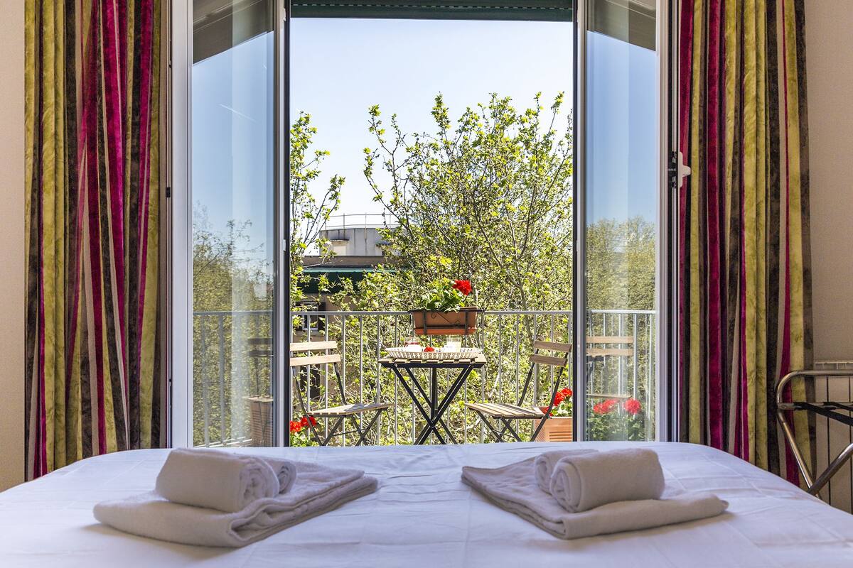 A hotel room with a bed, two towels, a balcony with a table and chairs, and a view of trees and buildings.