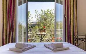 A hotel room with a bed, two towels, a balcony with a table and chairs, and a view of trees and buildings. - Top Floor Trastevere (Rome)