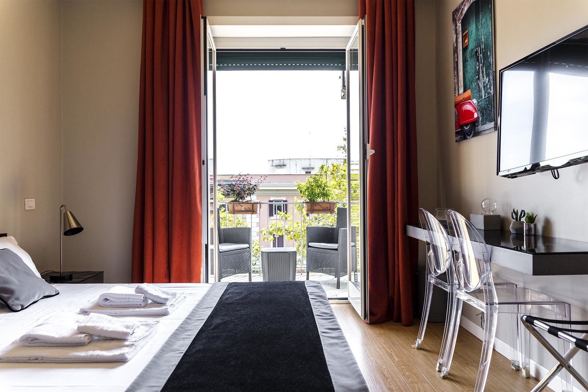 A hotel room with a bed, a desk, a chair, a TV, and a balcony with red curtains.