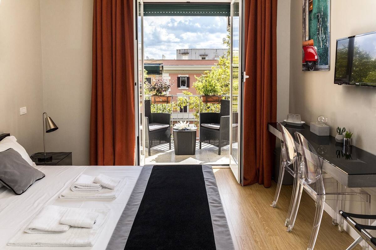 A hotel room with a bed, a desk, a chair, a TV, and a balcony with outdoor seating.
