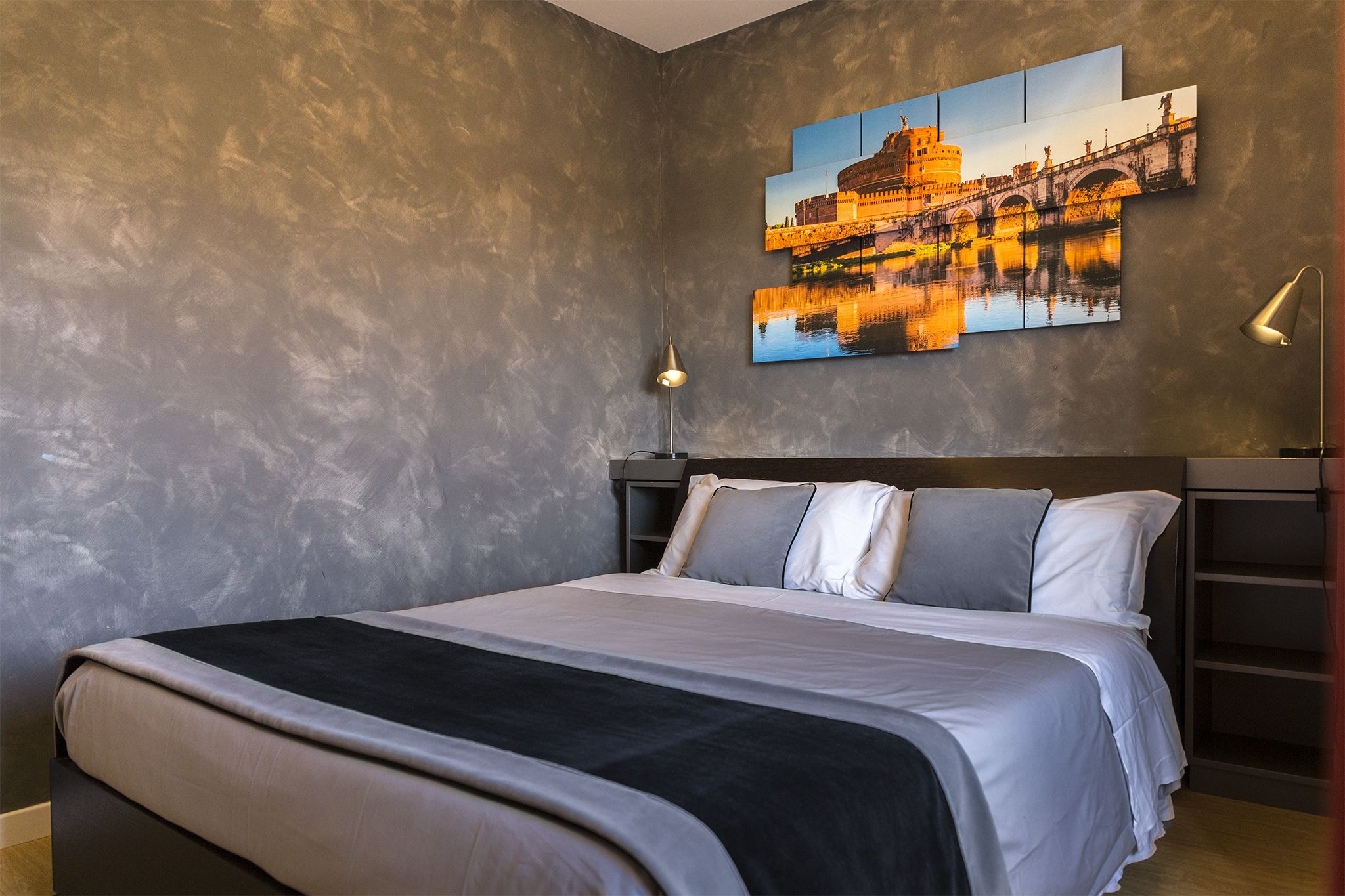A modern bedroom with a large bed, a nightstand, a wall-mounted lamp, and a cityscape painting.