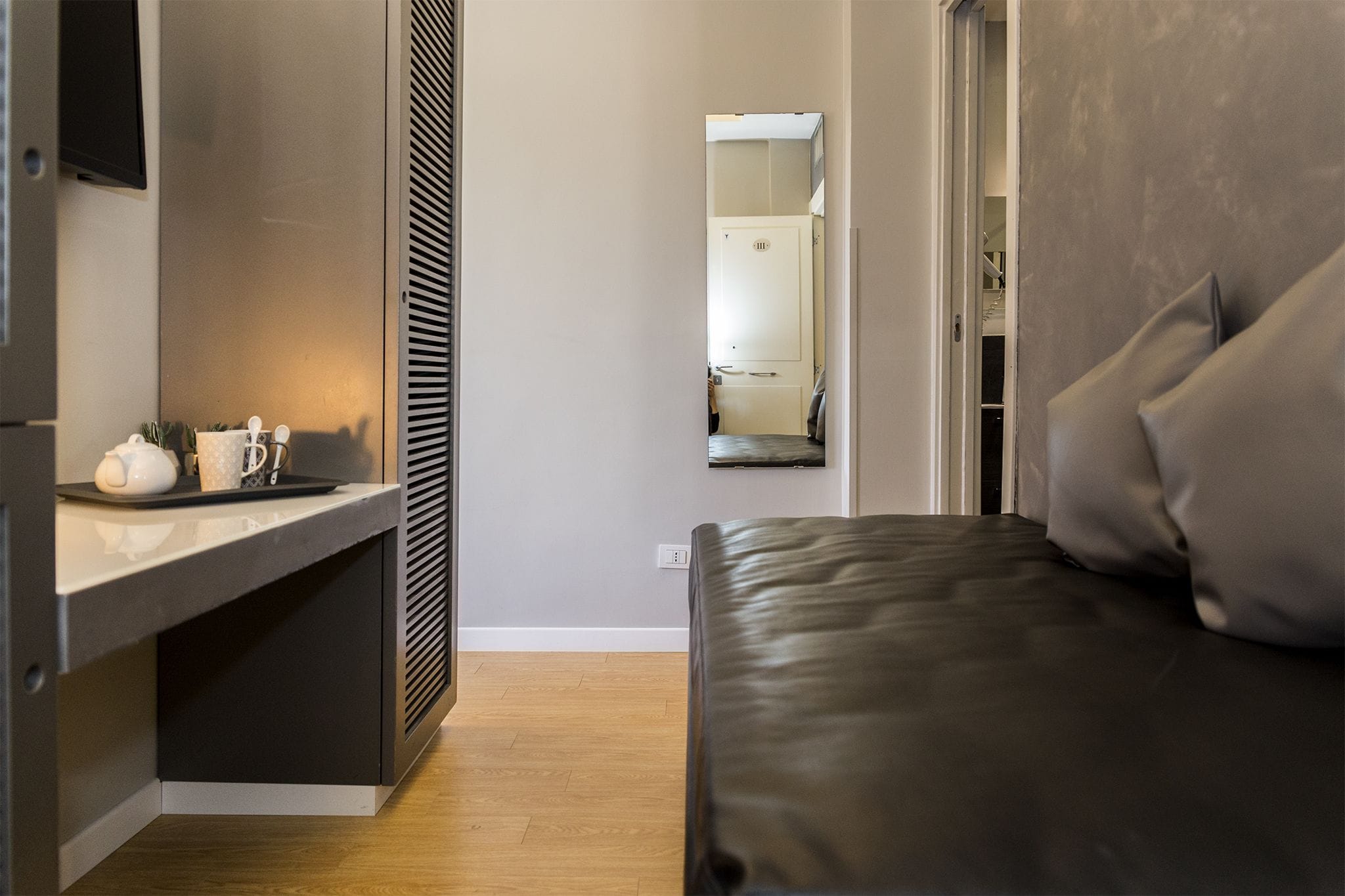 A modern room with a bed, a bedside table with a kettle and cups, a mirror, and a wardrobe.
