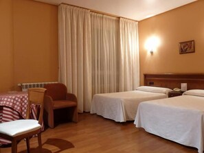 A hotel room with two single beds, a wooden nightstand, a chair, a table with a checkered tablecloth, and a large window with curtains. - Hotel Las Moreras (Sariegos)