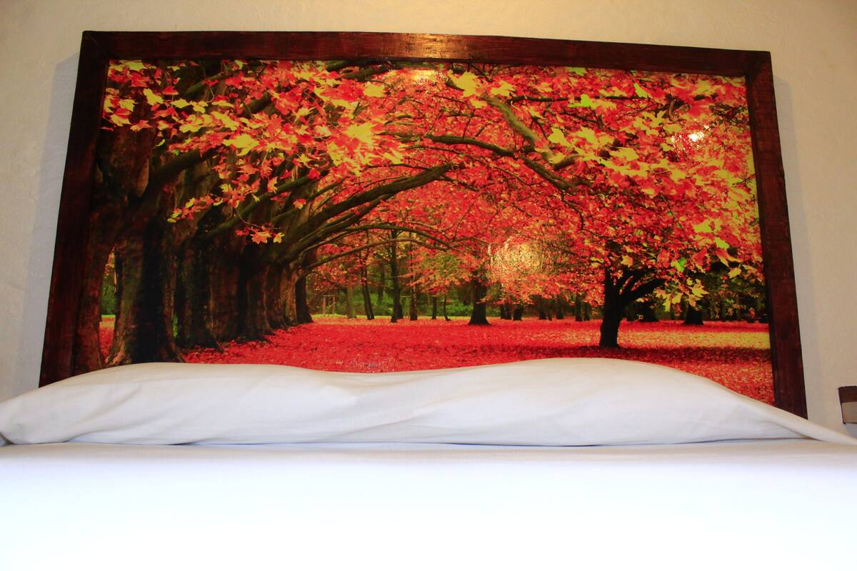 A bed with a white sheet and a large framed picture of a tree-lined path with red foliage.
