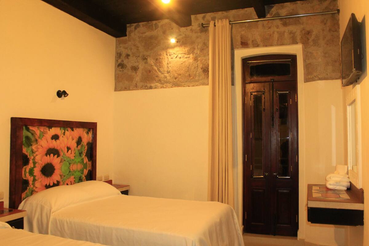 A hotel room with two beds, a large floral painting on the wall, a wooden door, a mirror, and a small table with towels.
