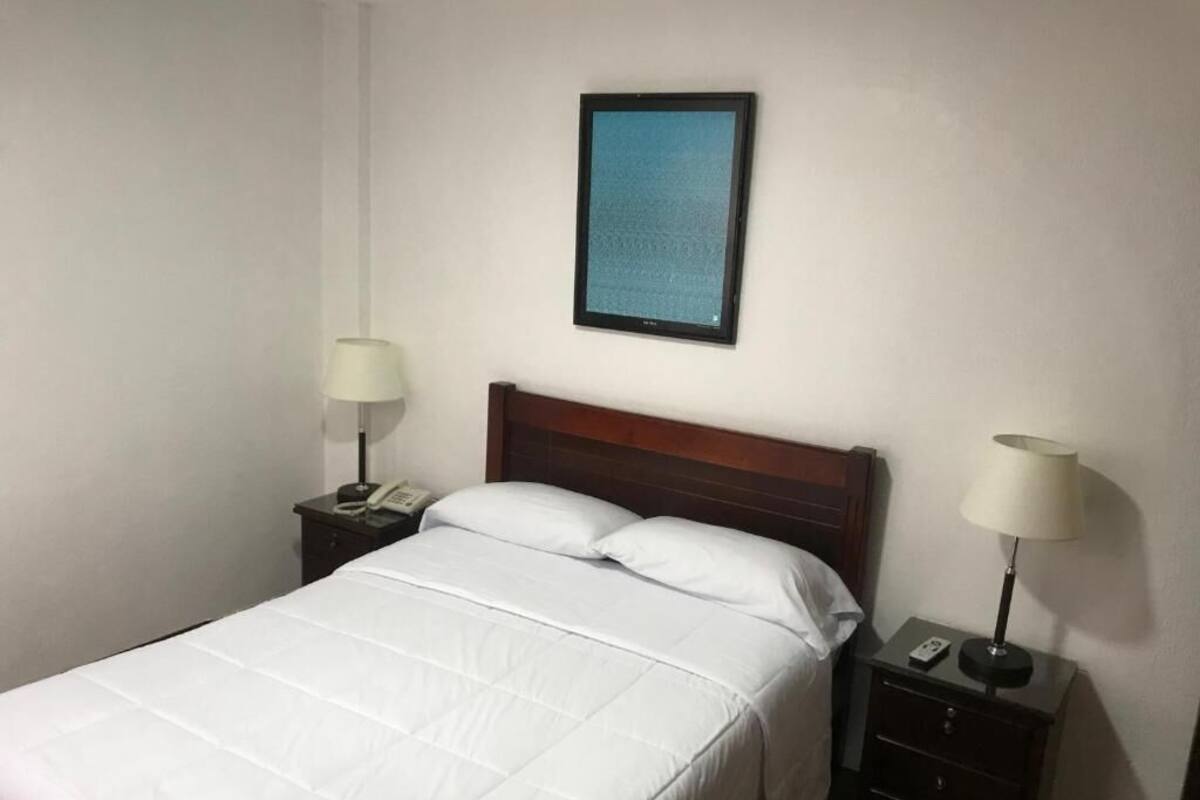 A neatly made bed with white bedding, a wooden headboard, and two bedside tables with lamps and a phone.