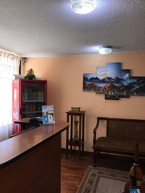 A reception area with a wooden desk, a Coca-Cola refrigerator, a small table with books, a sofa, and a wall with three paintings. - Hotel Altura (Quito)