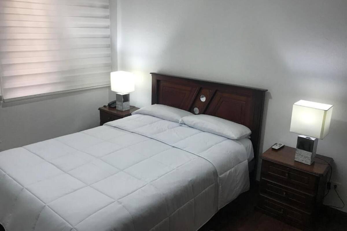 A neatly made bed with a white duvet, two bedside tables with lamps, and a window with blinds.