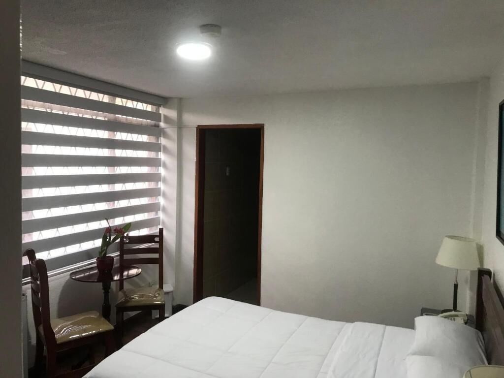 Quarto de hotel com cama, secretária com cadeira, abajur e janela com persianas.