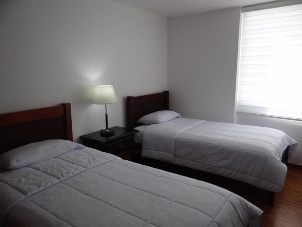 Una habitación con dos camas individuales, una mesita de noche con lámpara y una ventana con persianas.