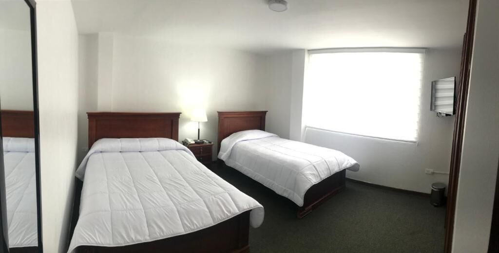 Una habitación de hotel con dos camas individuales, una mesita de noche con lámpara, una ventana con persianas y un cubo de basura.