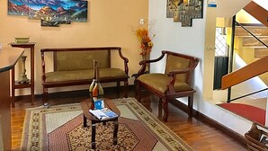 Een woonkamer met een bank, twee fauteuils, een salontafel en een vloerkleed. Op de muren hangen schilderijen en aan de rechterkant een trap.