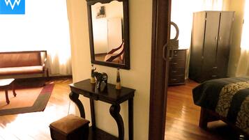 A room with a wooden floor, a bed with a patterned bedspread, a dark wooden desk with a mirror, a bench, and a wardrobe.