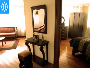 A room with a wooden floor, a bed with a patterned bedspread, a dark wooden desk with a mirror, a bench, and a wardrobe. - Hotel Altura (Quito)