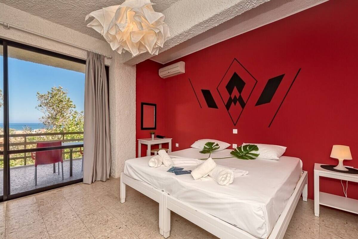 Habitación de hotel con una cama grande, vista al mar y una pared de acento roja.