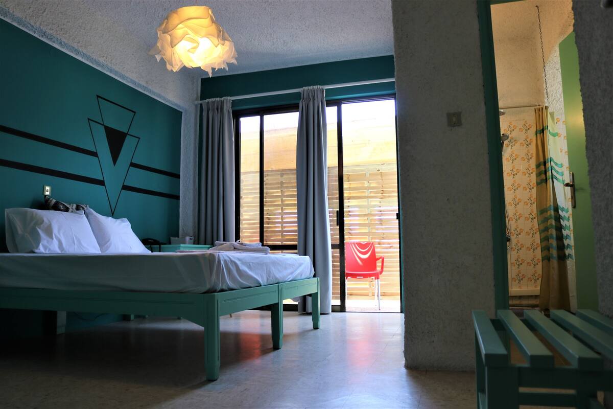 Un dormitorio con cama, un banco, una silla roja y vistas al exterior a través de un ventanal corredero.