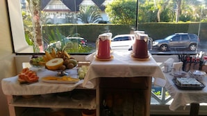 A breakfast buffet with various fruits, pastries, and beverages on a table near a window. - Pousada Bariloche (Teresópolis)