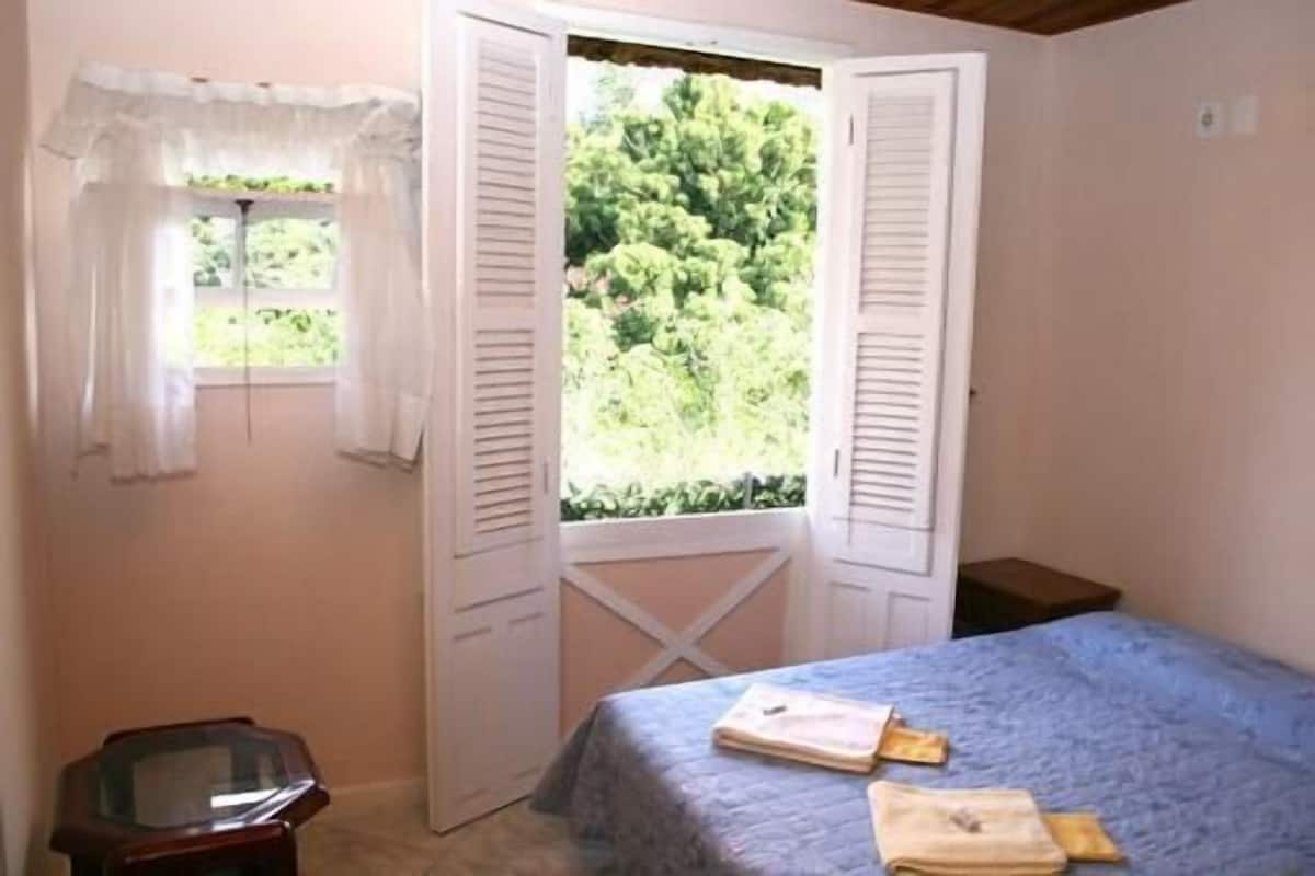 A bedroom with a bed, a window with open shutters, a small table, and a view of greenery outside.
