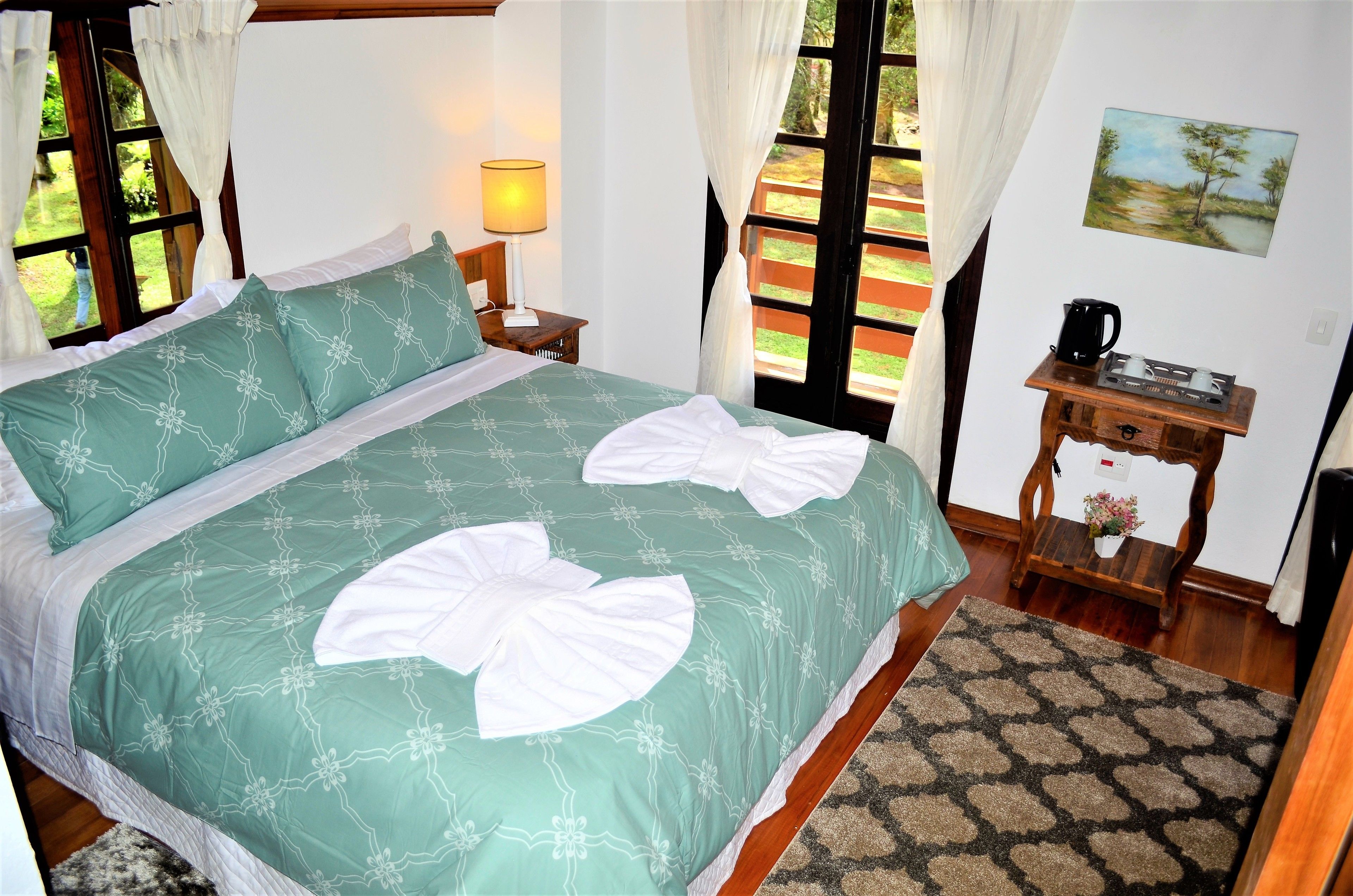 A neatly made bed with a green bedspread and white pillows, a bedside table with a lamp, a small wooden table with a coffee maker, and a painting on the wall.