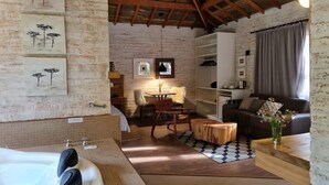 Una habitación rústica con techo de madera, pared de ladrillo blanco, un ventanal con cortinas, un sofá, una mesita de centro, una pequeña mesa de madera y una estantería de pared.