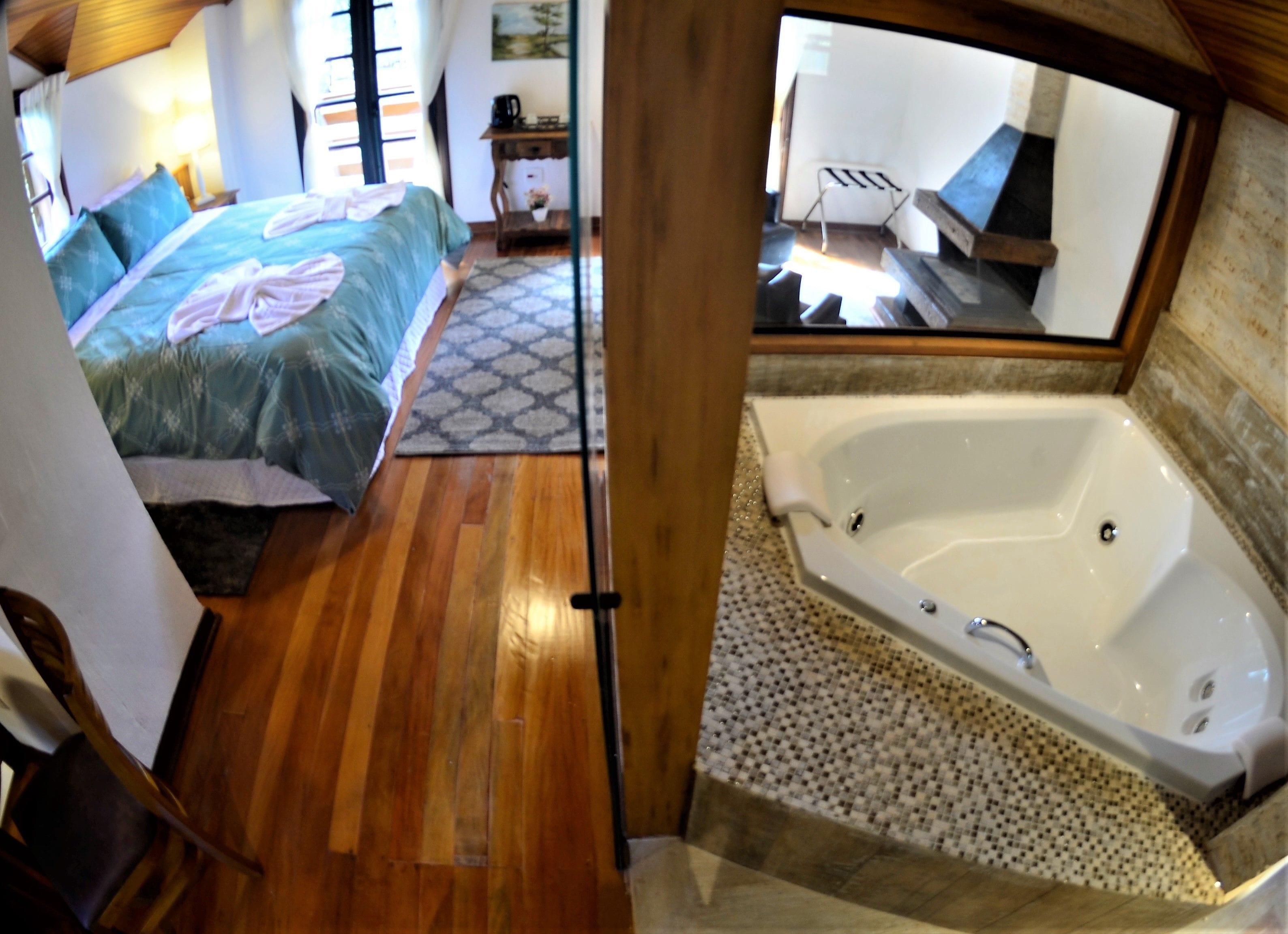 A bedroom with a large bed, a wooden floor, a bathroom with a bathtub, and a mirror reflecting the room.