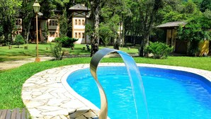 Una piscina con una fuente acuática curva, rodeada de baldosas de piedra y follaje.