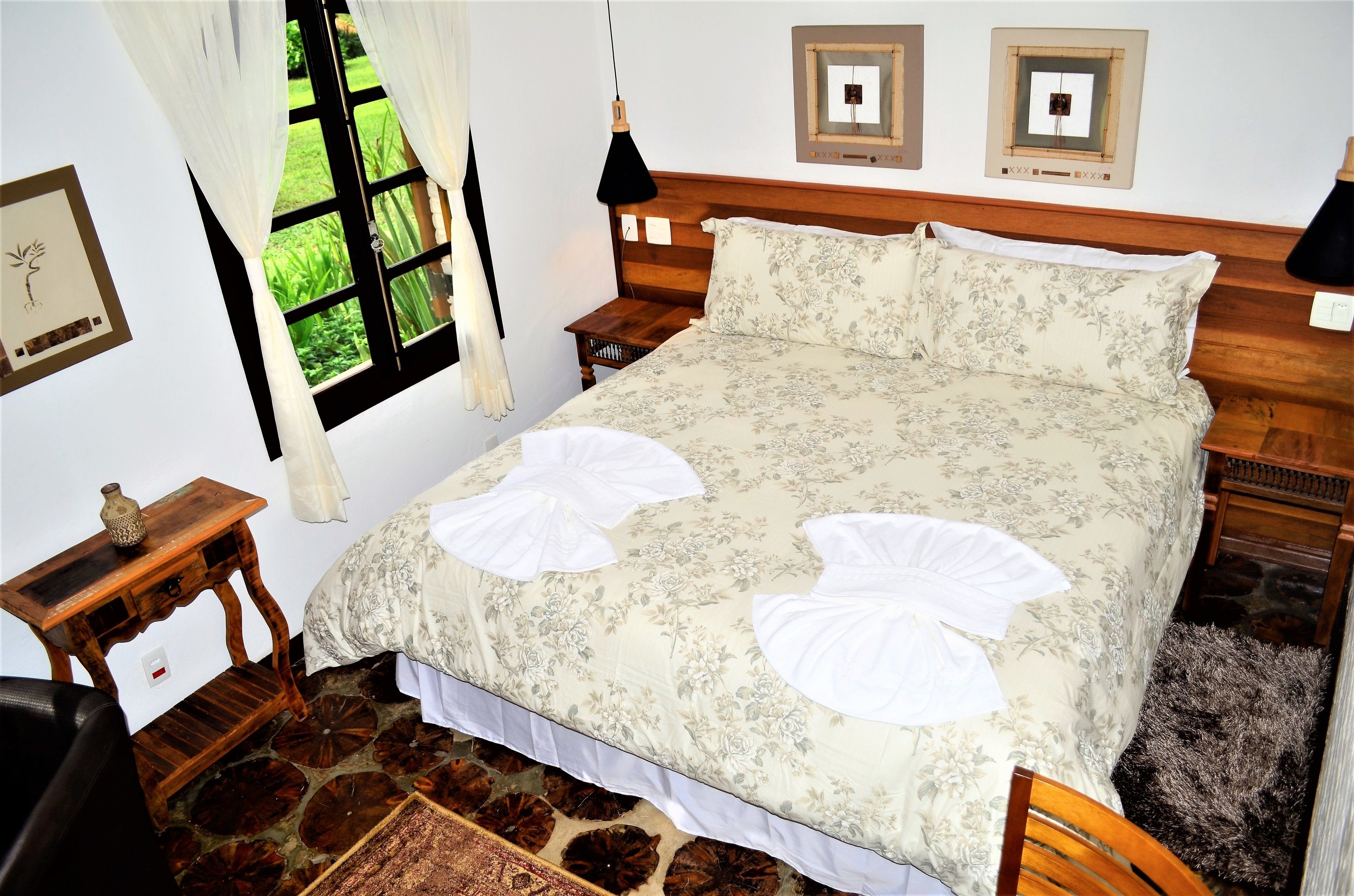 A bedroom with a wooden headboard, floral bedding, a nightstand, a bedside lamp, and a window with curtains.