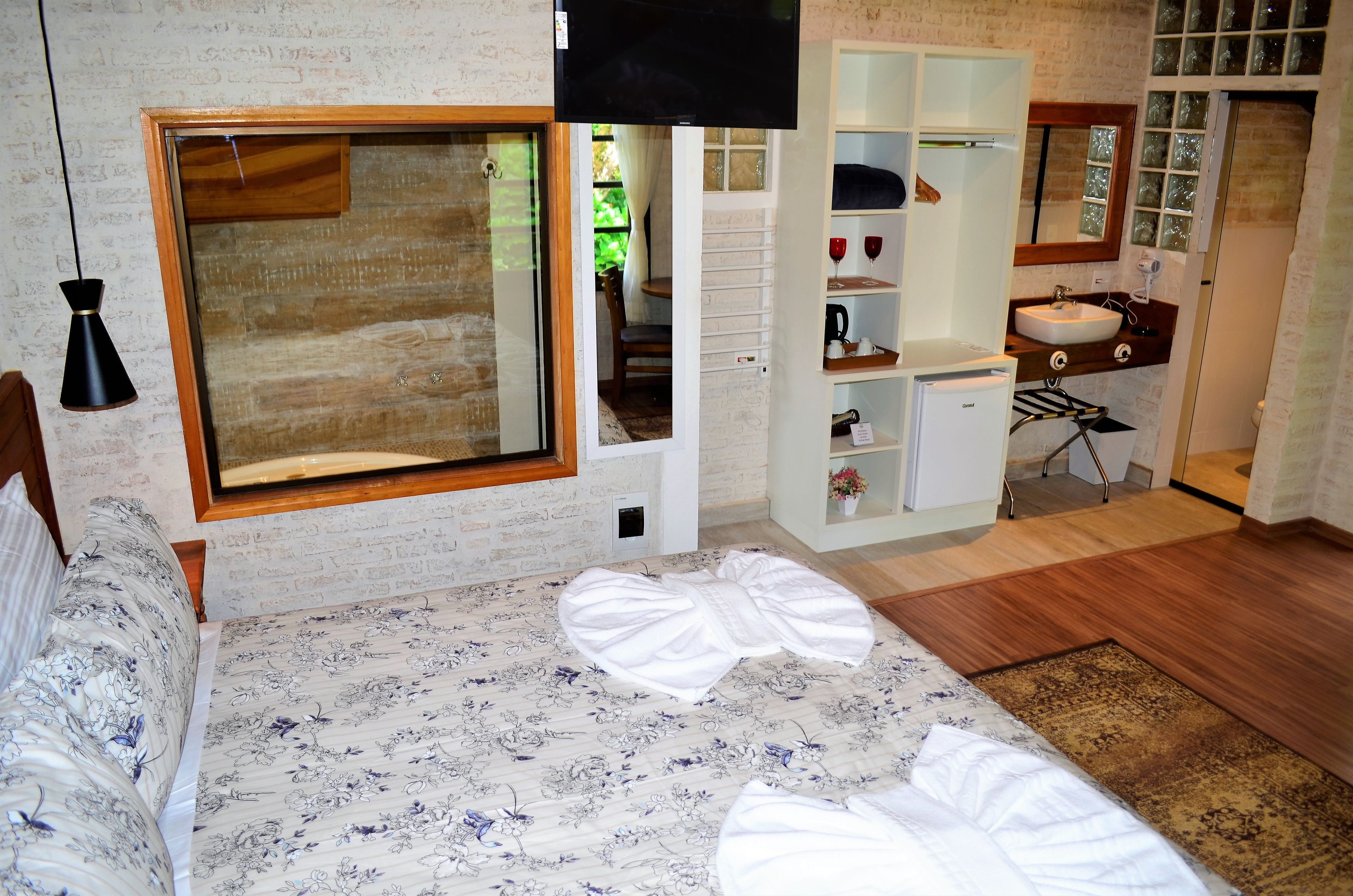 A bedroom with a large bed, a wooden-framed window, a built-in wardrobe, a bathroom with a sink and mirror, and a small desk.