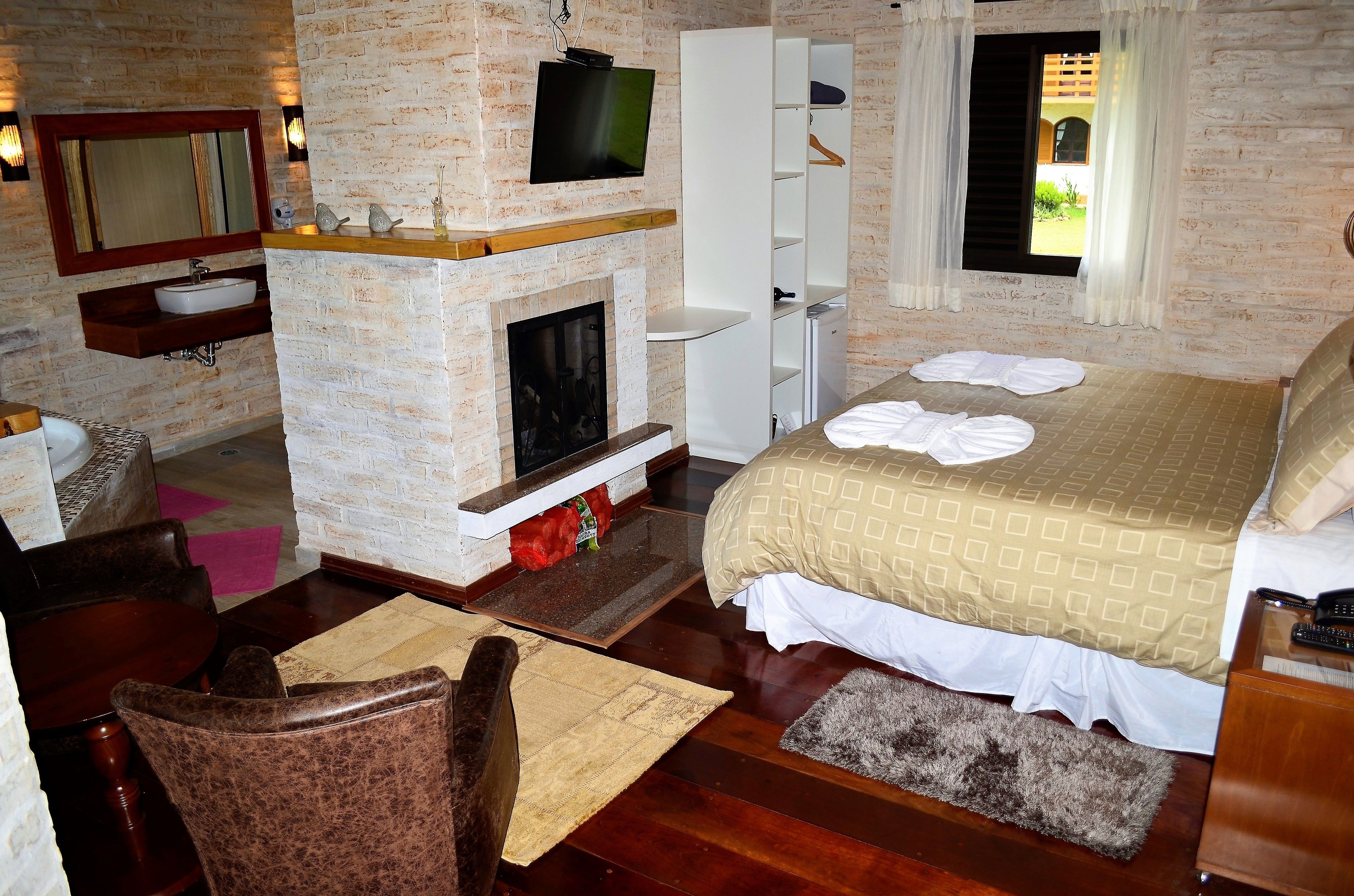 Habitación de hotel con pared de ladrillo, chimenea, cama con colcha estampada, televisor montado sobre la chimenea y un pequeño estante de madera.