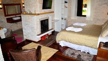Habitación de hotel con pared de ladrillo, chimenea, cama con colcha estampada, televisor montado sobre la chimenea y un pequeño estante de madera.