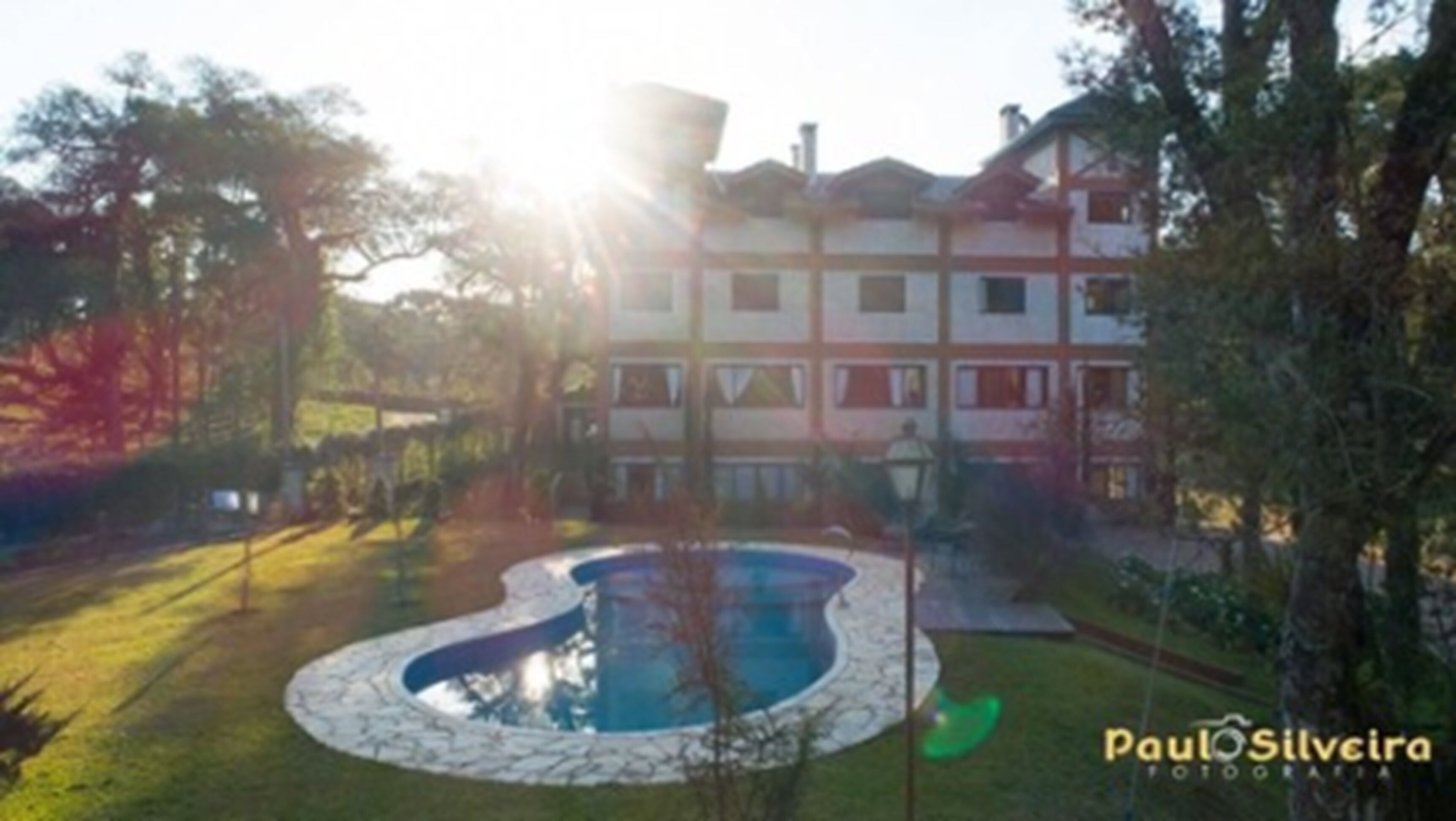 A building with a swimming pool surrounded by a stone border, trees, and a clear sky.