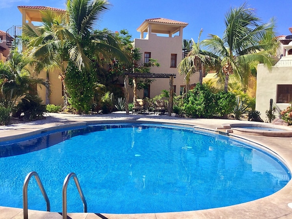 A swimming pool with a clear blue water, surrounded by palm trees and a building with a tiled roof.