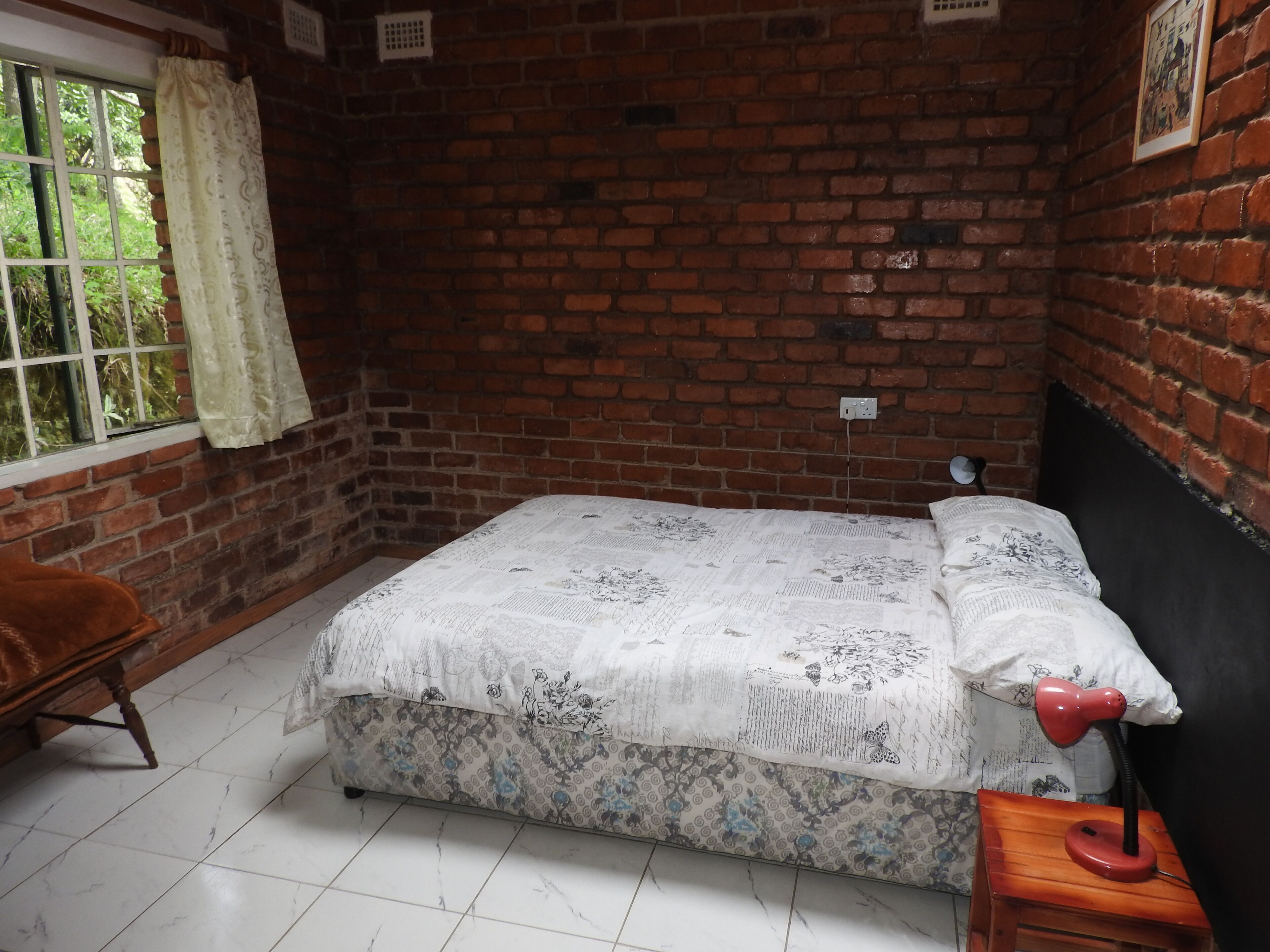 A room with a brick wall, a bed with a floral bedspread, a wooden nightstand with a red lamp, and a window with a curtain.