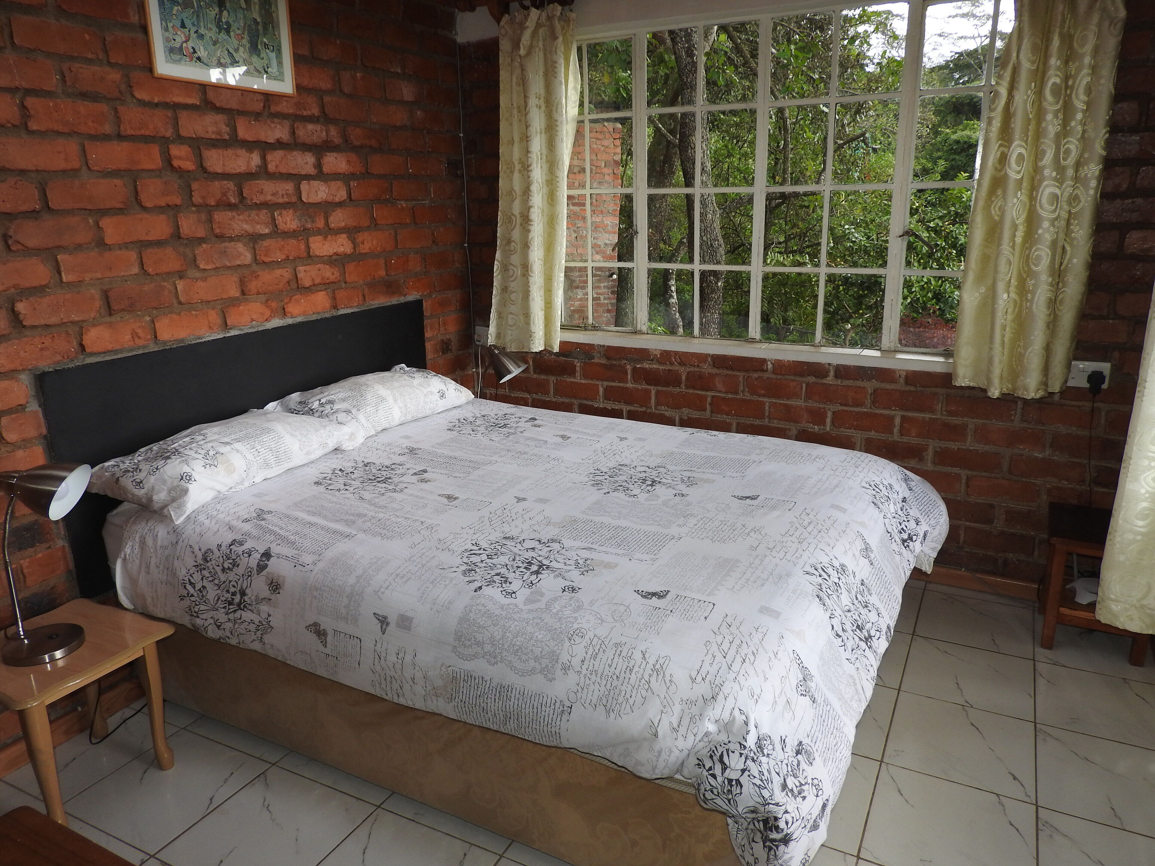 A bedroom with a brick wall, a bed with floral bedding, a nightstand, a desk, a window with curtains, and a view of greenery outside.