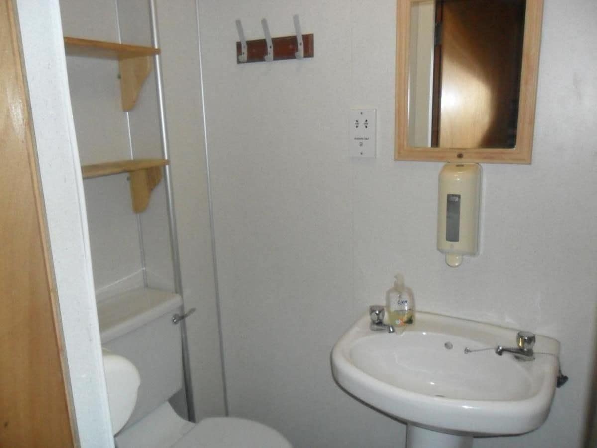 Un baño pequeño con inodoro, lavamanos y espejo. Hay estantes de madera sobre el inodoro y un dispensador de jabón en el lavamanos.
