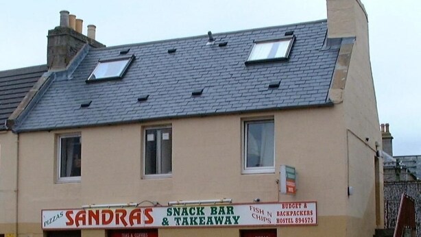 Un edificio con un letrero que indica 'Sandras Snack Bar & Takeaway', ofreciendo pizzas, tés, cafés y diversas comidas.