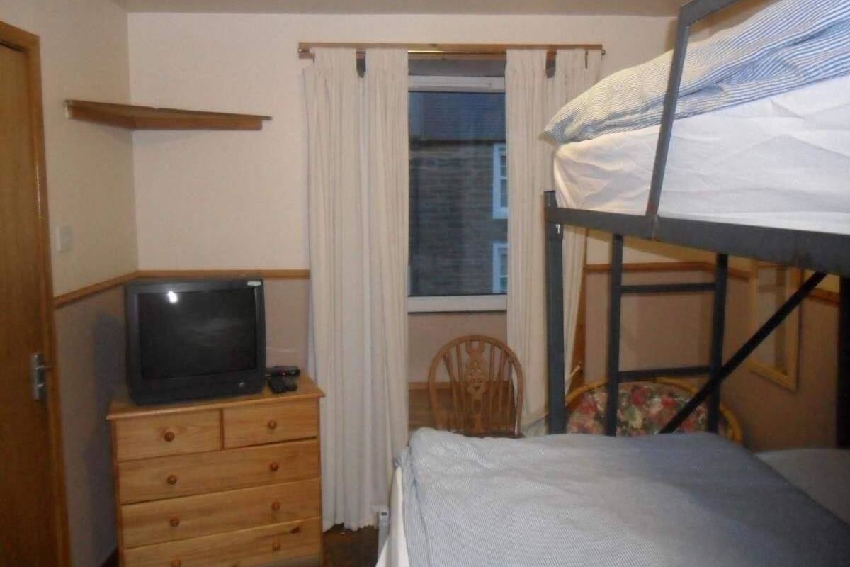 A small, compact room with a bunk bed, a wooden dresser with a television, a chair, and a window with curtains.