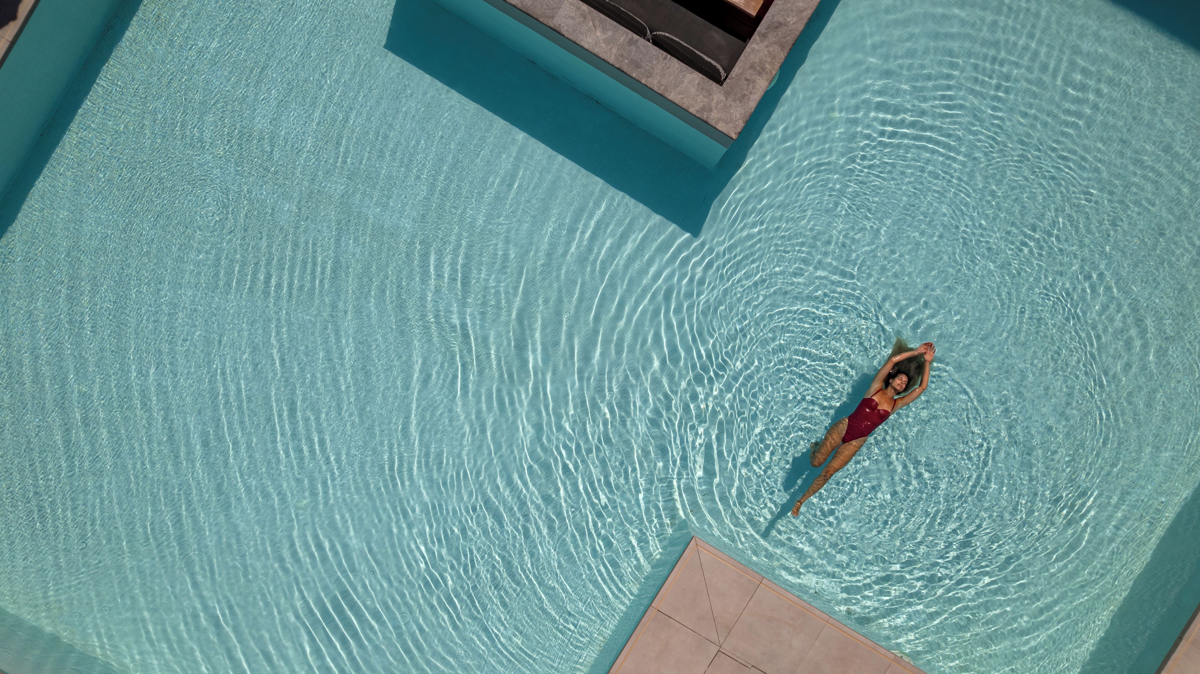 A person floating in a clear blue pool with a tiled deck area.