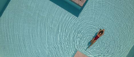 A person floating in a clear blue pool with a tiled deck area.